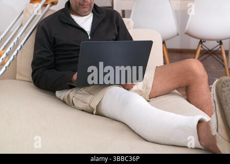 Man with broken leg working from home using laptop while sitting on couch with crutches nearby Stock Photo