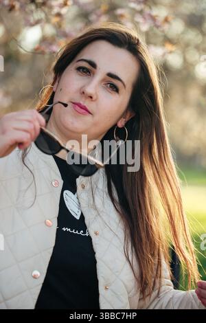 Young woman poses confidently outdoors wearing sunglasses during a sunny day in a park with blooming trees Stock Photo
