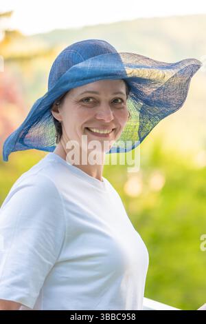 Middle-aged woman in white T-shirt and stylish summer hat smiling in nature Stock Photo
