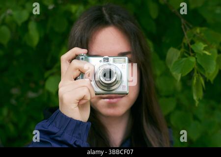 Girl holding old Canon compact digital camera, close-up against green foliage background Stock Photo