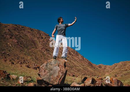 Handsome young man stands on big stone on mountains background and looks at his hand Stock Photo