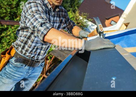 A carpenter diligently installs metal roofing on a residential building. The worker wears protective gloves and a plaid shirt, focusing on precision w Stock Photo