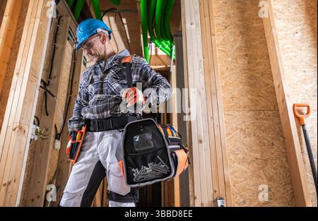 A construction worker is seen inside a partially finished building, wearing a hard hat and safety gloves. He is adjusting his tool belt and looking in Stock Photo