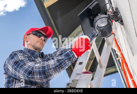A technician is working on a ladder to install a security camera on the exterior wall of a residential building. The day is bright with blue skies vis Stock Photo