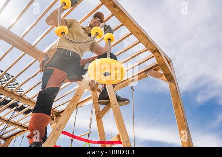 A man works intently on building a wooden playground structure outdoors. He is positioned on a ladder, carefully installing yellow rope grips. The set Stock Photo