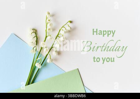Lily of the valley flowers arranged with pastel blue and green envelopes on a white surface. Text "Happy Birthday to you" in green adds a celebratory Stock Photo