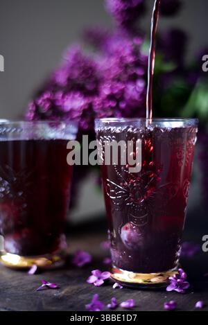 Elegant purple drink being poured into embossed glasses with lilac flowers in the background. Dark moody composition with floral elements. Stock Photo
