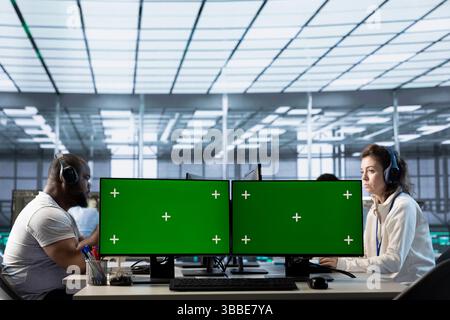 Technicians in data center listening music, optimizing IT infrastructure using green screen PCs. Server room experts wearing headphones upgrading gear with chroma key computers, reducing costs Stock Photo