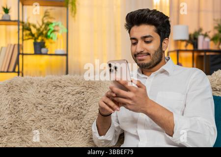 Indian young man at home sitting on sofa focused on smartphone typing text, browsing news, reacting to notifications. Arabian guy using mobile phone for social media, work, texting, digital lifestyle Stock Photo