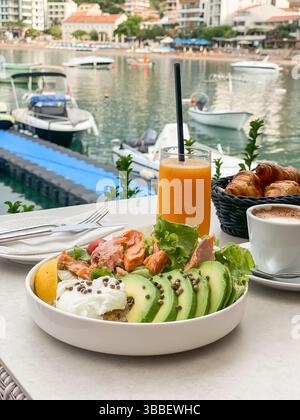 Healthy breakfast with avocado, poached egg, salad and baked salmon, coffee, orange juice and croissants in a waterfront cafe Stock Photo
