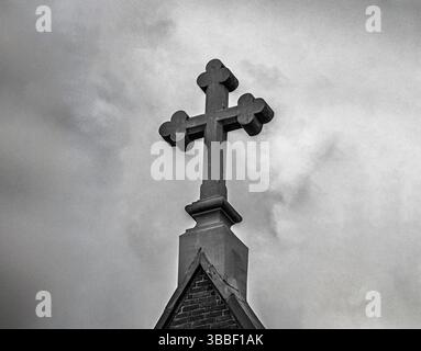 A dramatic black and white photo of a stone cross atop a church steeple, silhouetted against a moody, cloud-filled sky. Stock Photo