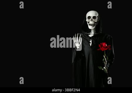 Scary human skeleton in death costume holding rose flower on black background Stock Photo