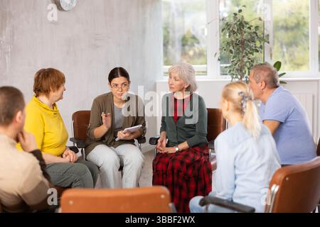 Young woman presides over support group meeting with mature people listening Stock Photo