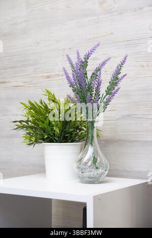 Minimalist indoor setup with artificial lavender in a glass vase and green plant in a white pot on a shelf. Stock Photo