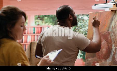 Female painter takes notes and teaches the art of painting in atelier, analyzing creative technique of her apprentice. Artist helping man to create nature inspired paintwork in workshop. Camera A. Stock Photo