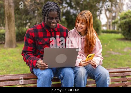 Two happy young students are using a laptop and a smartphone while sitting on a bench in a park, enjoying online learning and connectivity Stock Photo