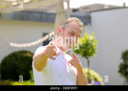 A smiling senior man in a white T-shirt points playfully at the camera with both hands. Stock Photo