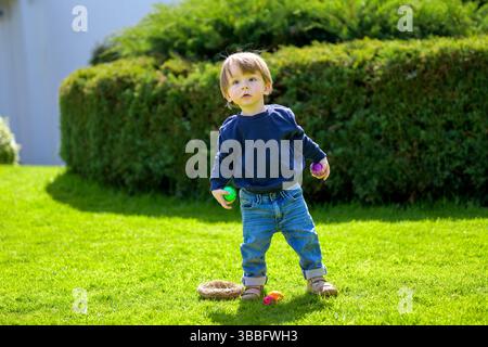 A toddler stands on a vibrant green lawn, holding colorful Easter eggs in both hands. Stock Photo