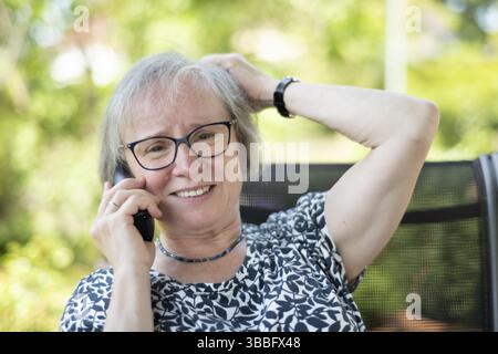 Outdoor shot, elderly woman, glasses, dressed in summer clothes, sitting in the garden, talking on the phone, smartphone, smiling, friendly, relaxed, Stock Photo