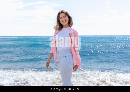 Smiling young woman looking at camera while standing on beach near ocean waves Stock Photo