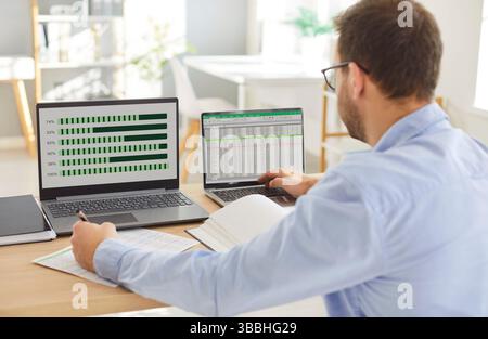 Man working at desk with two laptops with charts and spreadsheets, analyzing business data in office Stock Photo