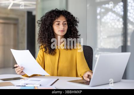A young woman in a yellow blouse sits at a desk, working on a laptop and reviewing documents. She is focused and engaged in her work. Stock Photo