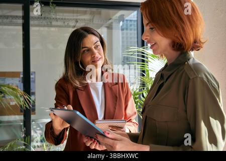 Two happy smiling business women talking using tab standing in green office. Stock Photo