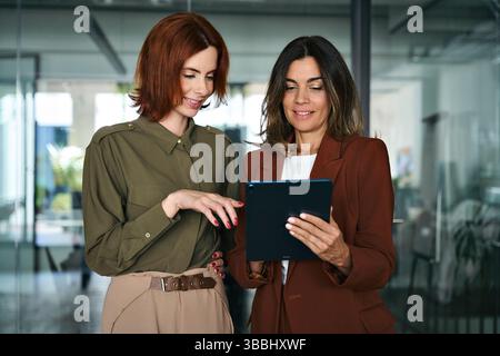 Two busy happy business women talking using tab working standing in office. Stock Photo