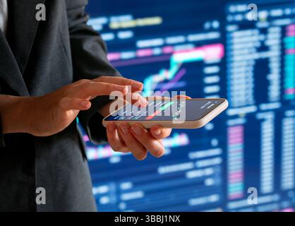 Female investor using smartphone for crypto trading in front of electronic chart Stock Photo