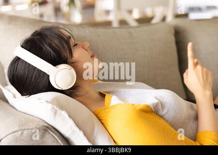 Listening Asian woman reclining on couch in living room, using smartphone with headphones Stock Photo