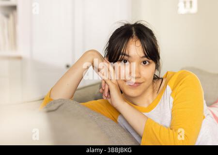 Reclining Asian woman supporting head with hands on light gray couch at home, with gray cushion Stock Photo