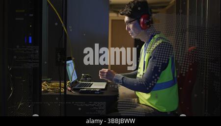 Operating laptop, technician wearing neon vest red earmuffs in server room with racks, copy space Stock Photo