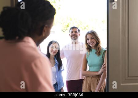 Diverse friends standing on sunlit porch, greeting host opening front door handle Stock Photo