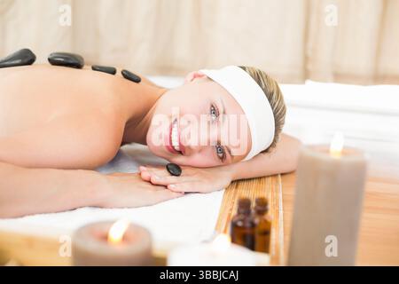 Relaxing female client lying on massage table in spa treatment room, with hot stones and candles Stock Photo
