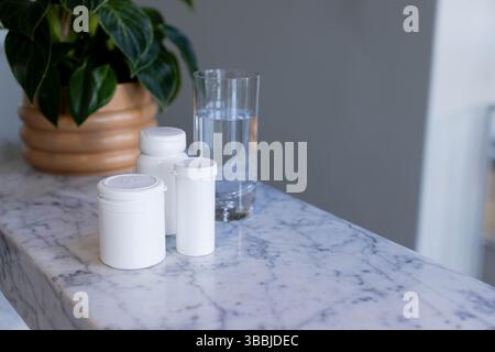 Three white medicine bottles are standing beside water glass on marble countertop by terracotta pot Stock Photo