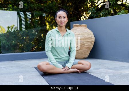 Practicing meditation Asian woman sitting cross-legged on yoga mat on stone terrace, with woven urn Stock Photo