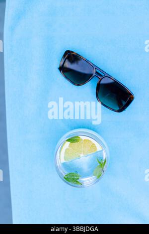 Glass of water with lemon slice and mint leaves sitting on blue towel, dark sunglasses resting Stock Photo