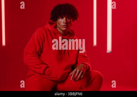 Posing man wearing red hoodie and sweatpants sitting in red studio, with neon tube lights Stock Photo