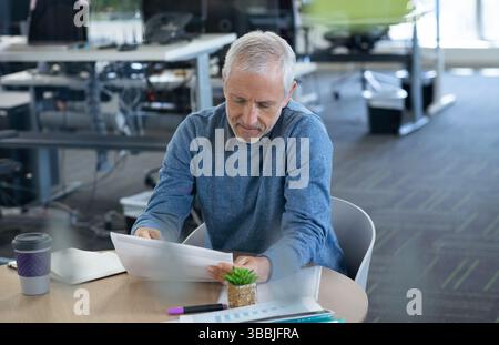Reading senior man reviewing documents at table in office, with coffee cup and succulent Stock Photo