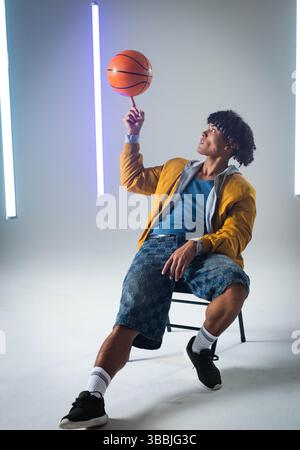 Man in bomber jacket spinning basketball on finger while sitting in studio, with vertical LED tubes Stock Photo