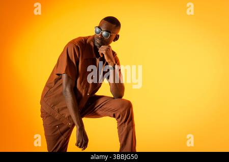 Leaning African American man on one knee in studio orange backdrop, with sunglasses, smartwatch Stock Photo