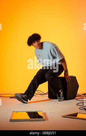 Sitting adult male model wearing textured sweater on speaker box in studio, with mirrors and cables Stock Photo