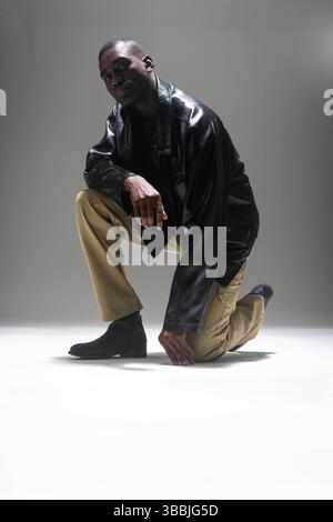 African American man kneeling on floor in studio, wearing leather jacket, cargo pants and boots Stock Photo