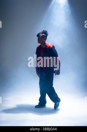 Standing African American male wearing black and red jacket on smoky stage, with spotlight beam Stock Photo
