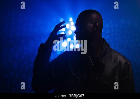 Standing African American man raising hand near face against dark stage, with necklace, blue lights Stock Photo