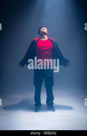 Posing African American man wearing red top and black jacket with chain in studio under spotlight Stock Photo
