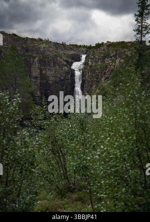 Njupeskär Waterfall flows dramatically down a rugged cliff in Fulufjället National Park, surrounded by dense foliage and under a moody, overcast sky Stock Photo
