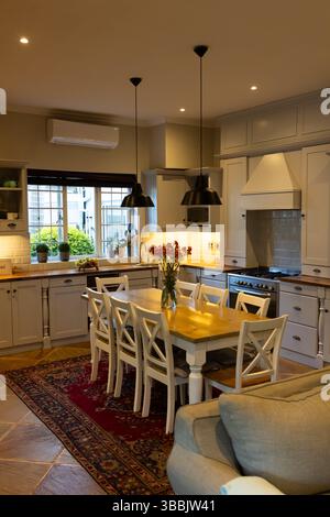 Wooden dining table standing in home kitchen with white chairs, black pendant lamps, potted plants Stock Photo