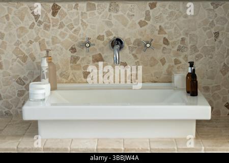 White sink basin is sitting on stone countertop, displaying chrome faucet and cosmetic bottles Stock Photo