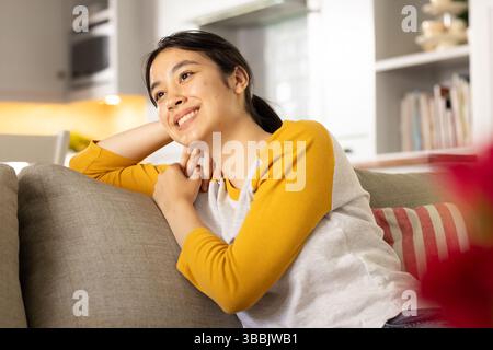 Leaning Asian woman resting head on gray sofa in living room, with red-and-white striped cushion Stock Photo